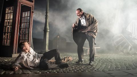 Two men on the set of An Inspector Calls. The younger man lays on the floor as an older man stands over him. Smoke fills the air behind them.