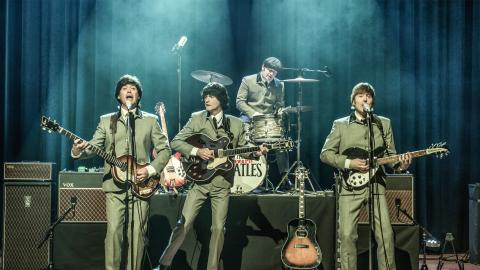 Four members of tribute group the Cavern Beatles stand on stage playing guitars and drums.