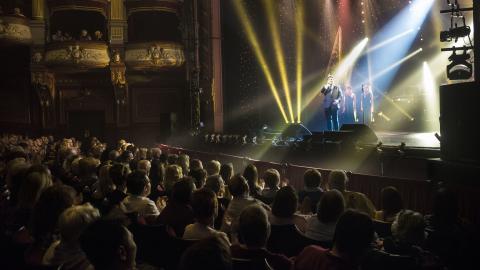 Audience sat watching Michael Ball performing on stage at the Alhambra Theatre with yellow stage lights shining upward.