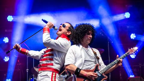 Tribute artists of Freddie Mercury and Brian May stand performing on stage holding a microphone and guitar. 