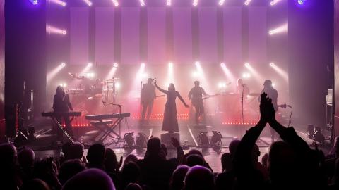 Five silhouettes on stage of tribute band Rumours of Fleetwood Mac, with pink and purple lights behind them. 