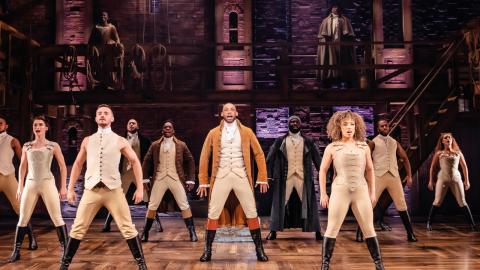 Production Image of Hamilton, cast members stood on stage in neutral coloured clothing, with their hands to their side.