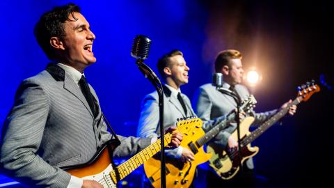 Three members of Rave On playing guitar and singing into microphones against a blue background.