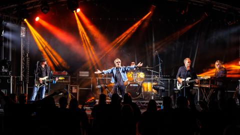 The Bon Jovi Experience production image with a group of performers on stage, red and orange stage lights shine down on them.