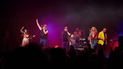 A group of musicians perform on stage against purple and red stage lights.