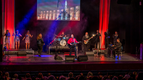 A group of musicians on stage singing and playing instruments with red curtains behind them and lights shining on the stage.