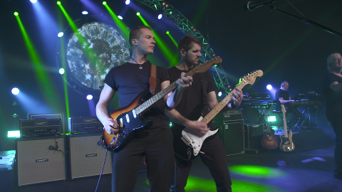 Two musicians on stage playing guitar against blue and green stage lights.