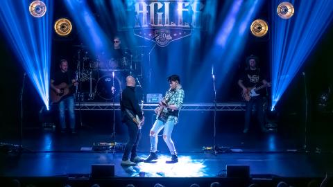 Musicians performing on stage together, playing guitars with blue and white stage lighting shining down on them.