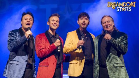 4 men wearing multi-coloured blazers singing on stage