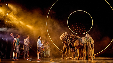 Production image of The Lion, The Witch and The Wardrobe with a group of children and a large puppet of a lion on stage surrounded by orange light.