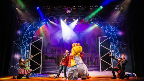 A set on stage of a concert with brightly coloured stage lights shining on a boy and someone in a dinosaur costume.