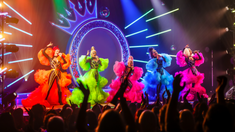 5 Drag queens performing on stage in black catsuits with brightly coloured, feather coats.
