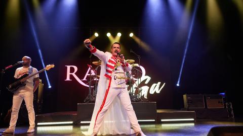 A Freddie Mercury tribute artist on stage wearing a red and white outfit, with blue and yellow stage lights shining down on them.