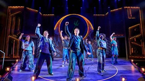 The cast of The Lightning Thief perform on stage surrounded by blue lights, all raising their right arm in the air.