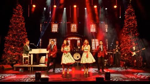 Four performers singing on stage wearing festive outfits, surrounded by red stage light.