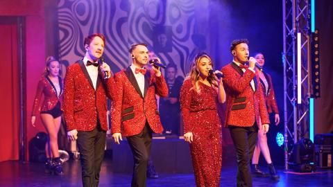 Four singers dressed in bright red outfits singing into microphones against a purple background.