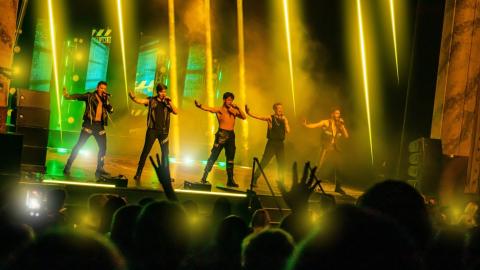5 men performing on stage wearing all black with yellow stage lights beaming down on them.