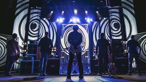 Musicians stood on stage waiting to perform with the lights off, screens in the black play a black and white swirling pattern.
