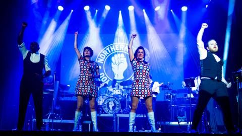 Four musicians on stage with blue stage lights behind them, raising their arms in the air.