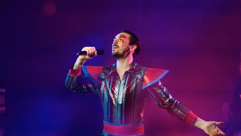A close up of a singer in a multi-coloured jump suit singing into a microphone.