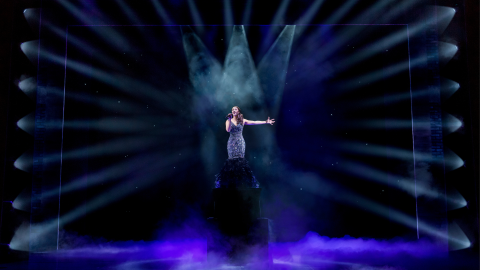 A woman singing in the centre of the stage dressed in silver, with white stage lights pointing at her, against a black background.