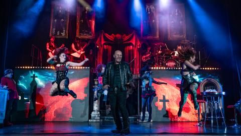 A man performing on stage wearing an all black, leather outfit with singers and musicians in the background.
