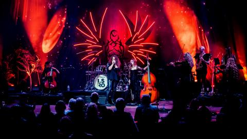 Musicians and singers performing on stage against red and orange dramatic lighting.
