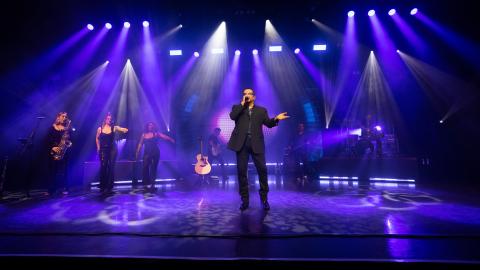 A George Michael tribute artist performing on stage with musicians in the background and purple stage lights.
