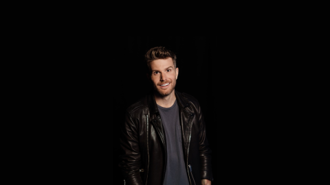 Comedian Joel Dommett stood against a black background wearing a black tshirt and leather jacket.