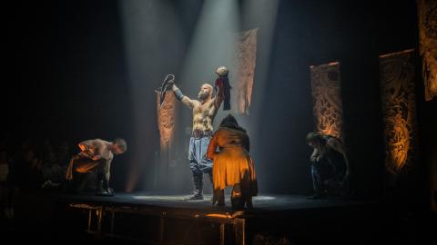 Performers on stage wearing Viking inspired clothing with dramatic white lighting shining down on them.