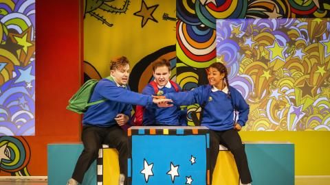 Three school students on stage surrounded by a bright background.