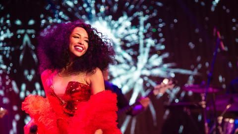 A woman in a long, red dress stands on stage against a projection screen.