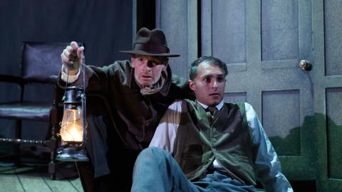 A man laying on the floor with another man leaning over him holding a lantern.