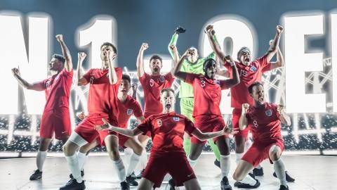 Group of cast members in Dear England wearing red England football kits, celebrating a goal.