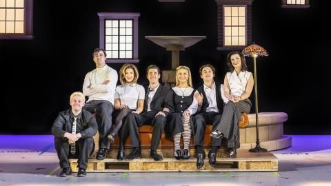 Seven cast members of Friends The Musical Parody sit on a sofa by a fountain, all wearing black and white outfits.