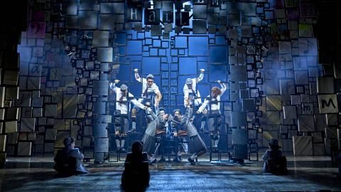 Cast members of Matilda The Musical climb gym apparatus on stage with white lights shining down on them.