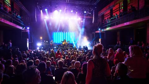 A crowd watching a musician sing and play instruments on stage, with bright stage lights.