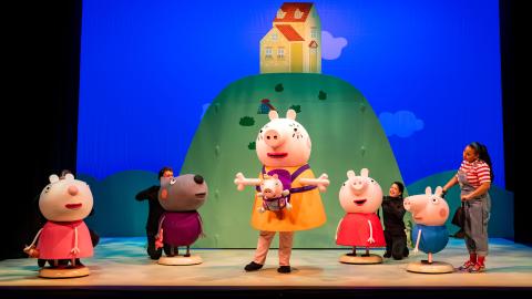 Characters of Peppa Pig on stage against a blue background and the house on a hill.