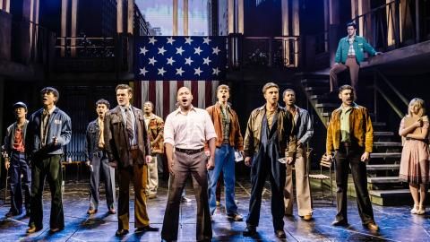 Cast members of Miss Saigon on stage looking forward, all wearing office or work wear.