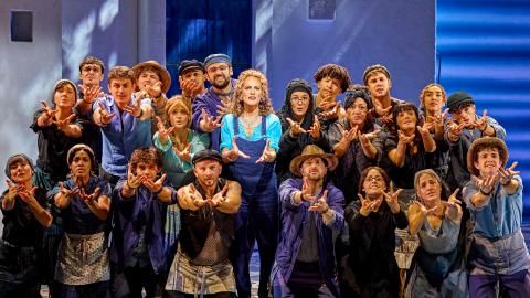 A woman wearing a blue shirt and dungarees holds her hands out, surrounded by dancers.