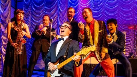A man kneels on the floor while singing and playing guitar, backing singers and musicians stand behind him.
