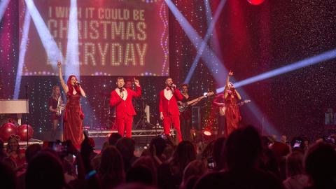 A festive live concert with singers in red outfits performing to a packed audience, arms raised under bright stage lights as artificial snow falls, with a large screen behind them reading “I Wish It Could Be Christmas Everyday.”