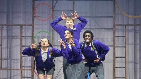 Actors performing as school children strike silly poses while stood in a school gym setting on stage.