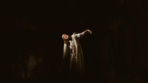 A woman in a white dress wearing a crown leans to the side with her arm in the air on a dark stage