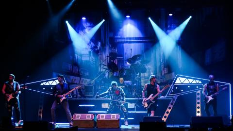Rock band performing onstage with dramatic blue lighting, featuring multiple guitarists and a drummer against a city‑scape backdrop.
