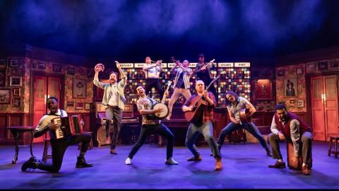 A group of male actors on stage in a bar setting, playing various instruments on stage.