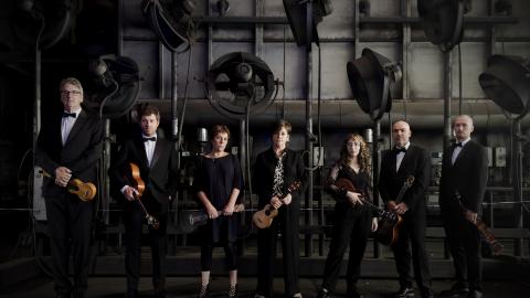 A group of musicians all wearing black, stood against a dramatic, dark setting.