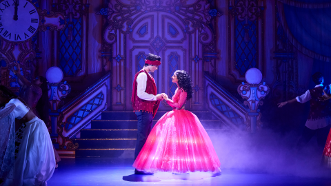 A woman, wearing a large pink dress holds hands with a man on stage in Surinderella.