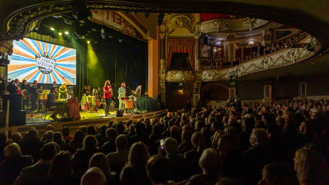 A group of musicians perform on stage with bright yellow lights behind them, in the foreground the audience face the stage.