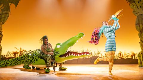 A woman operated a large crocodile puppet, sat against an orange backdrop. Another woman wears a bird outfit.